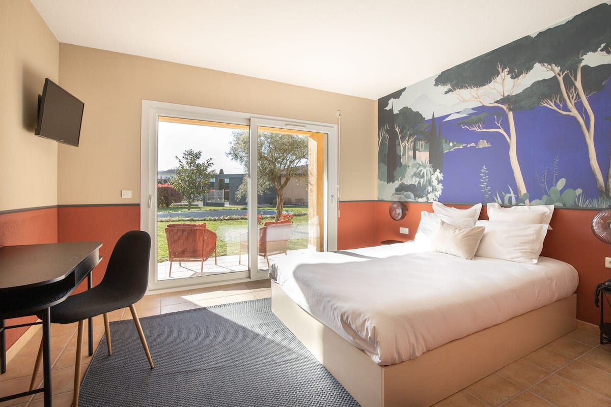 A bedroom with a bed, a desk, and a chair. There is a sliding glass door leading to an outdoor area with a view of trees and a building.