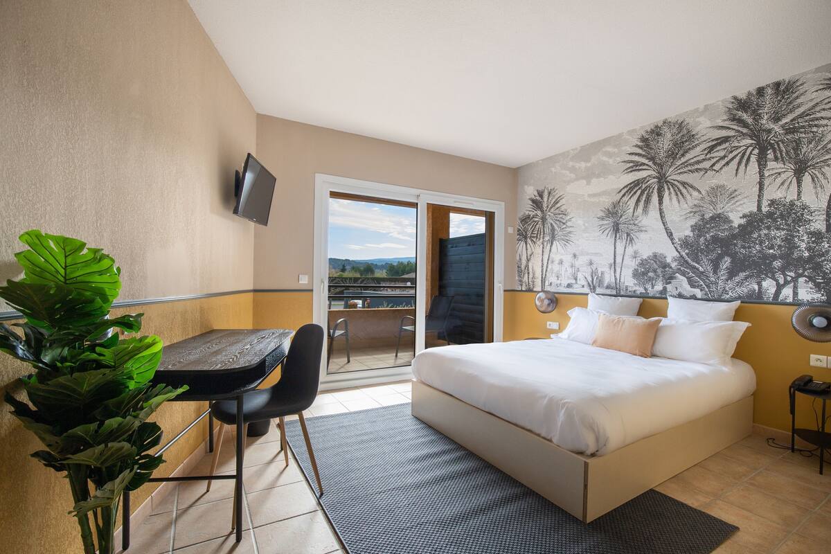 A hotel room with a bed, a desk, a chair, a TV, and a balcony with a view.