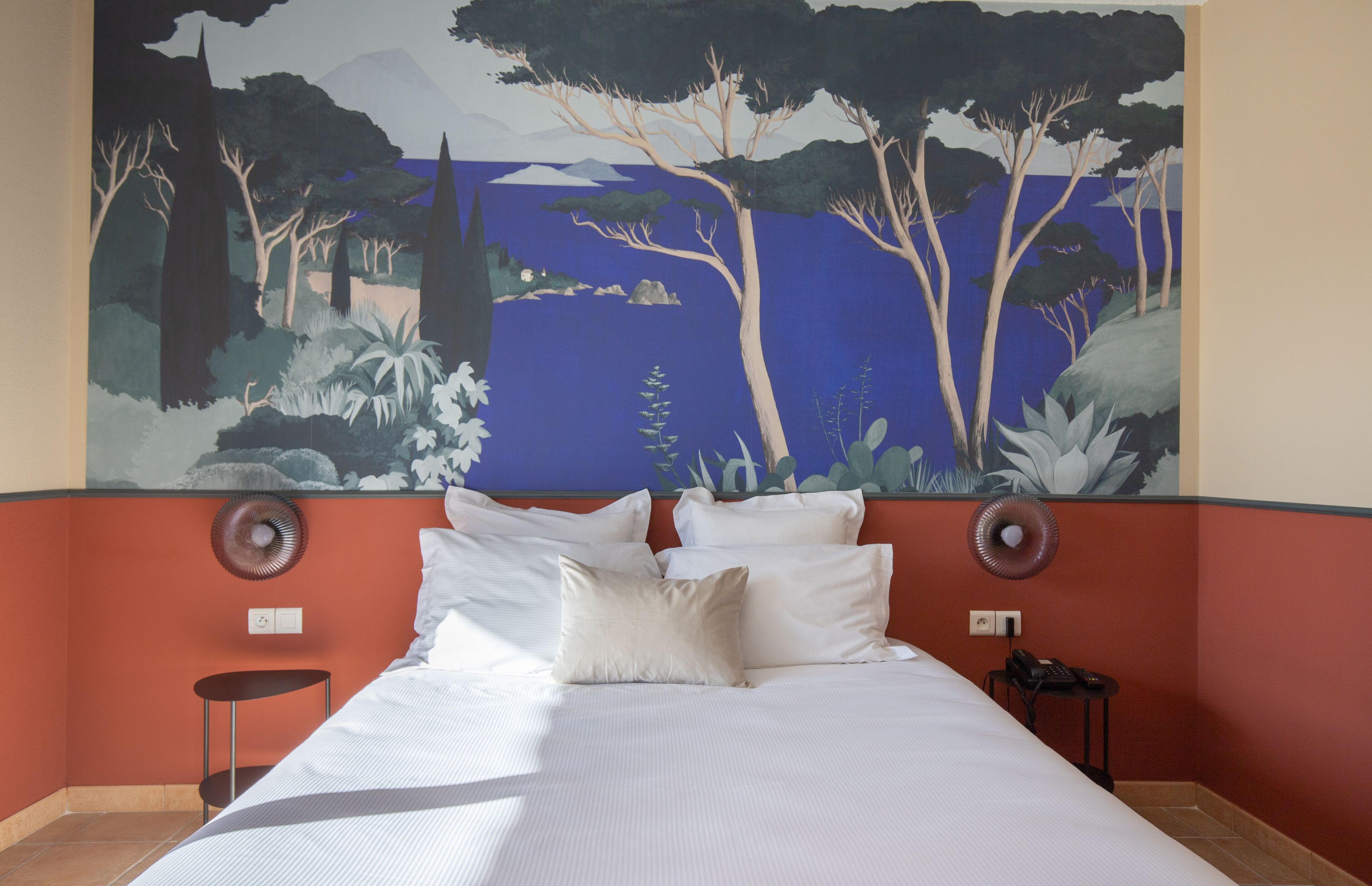Quarto de hotel com um grande mural de uma floresta e lago, uma cama com lençóis brancos, duas mesas de cabeceira e uma parede de destaque vermelha.