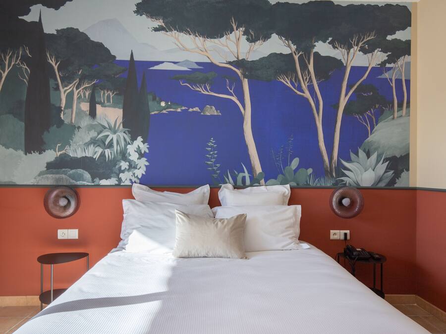 Quarto de hotel com um grande mural de uma floresta e lago, uma cama com lençóis brancos, duas mesas de cabeceira e uma parede de destaque vermelha.