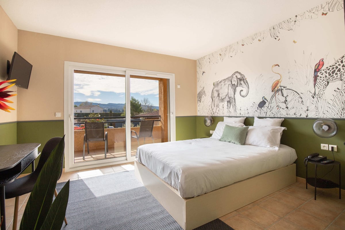 A bedroom with a bed, a desk, and a chair. There is a sliding glass door leading to an outdoor area with a table and chairs.