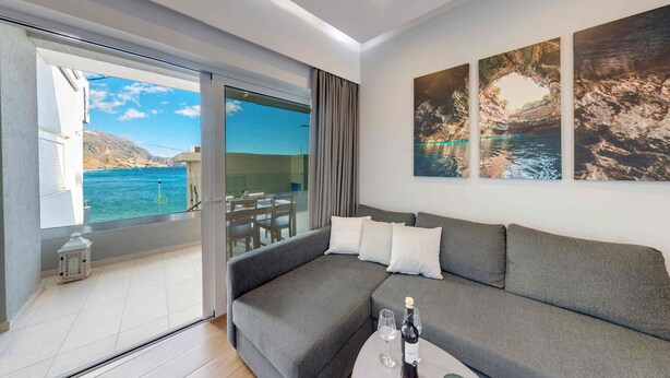 Een moderne woonkamer met een grijze bank, een glazen deur naar een balkon en drie ingekaderde schilderijen van kustlandschappen.