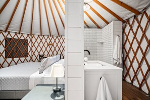 A modern, well-lit interior of a yurt with a bed, a small table with a lamp, and a bathroom with a sink and towel rack. - Ot 3515a Texas Yurt Haus: Armadillo 1 Bedroom Cabin by RedAwning (New Braunfels)
