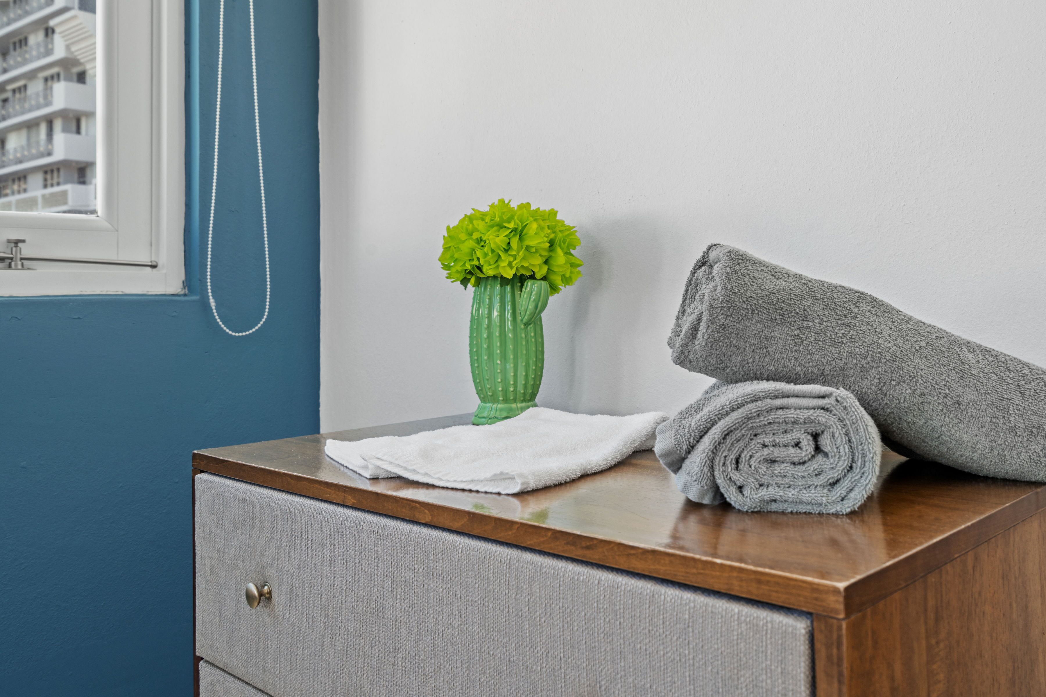 A bathroom with a wooden vanity, a green vase with flowers, a rolled towel, and a white towel on the counter.