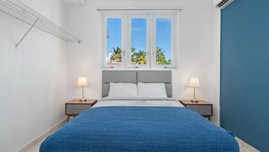 A bedroom with a bed, two bedside tables with lamps, a window with a view of palm trees, and a blue accent wall. - Puerto Rico 1 Bed Beach Rental Otium Rentals (San Juan)