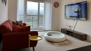 Un salon moderne avec un canapé rouge, une table basse ronde blanche, une petite table d’appoint jaune, un grand écran de télévision et un balcon avec vue.