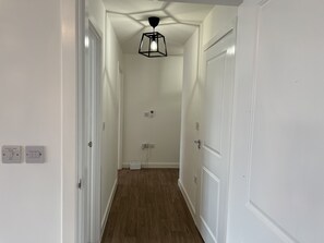 Un couloir étroit doté d’un plancher en bois, de murs blancs et d’un luminaire suspendu noir. De chaque côté, on trouve deux portes blanches, et des prises électriques sont visibles sur les murs.