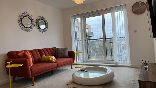 Un salon moderne avec un canapé rouge, une petite table d’appoint jaune, une table basse blanche et une grande fenêtre donnant sur des immeubles.