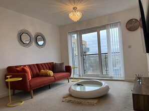 A modern living room with a red sofa, a small yellow side table, a white coffee table, and a large window with a view of buildings. - Mordern Retro 2 Bedroom Apartment (Erith)