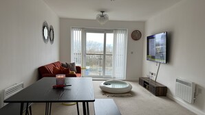 Un salon comprenant un canapé, une table avec des bancs, un téléviseur, un radiateur et une fenêtre avec des stores.