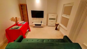 A compact living space with a green sofa, a dining table with red tablecloth, a flat-screen TV, a white cabinet, and a window with curtains. - Fully-equipped Flat in the City of London (London)