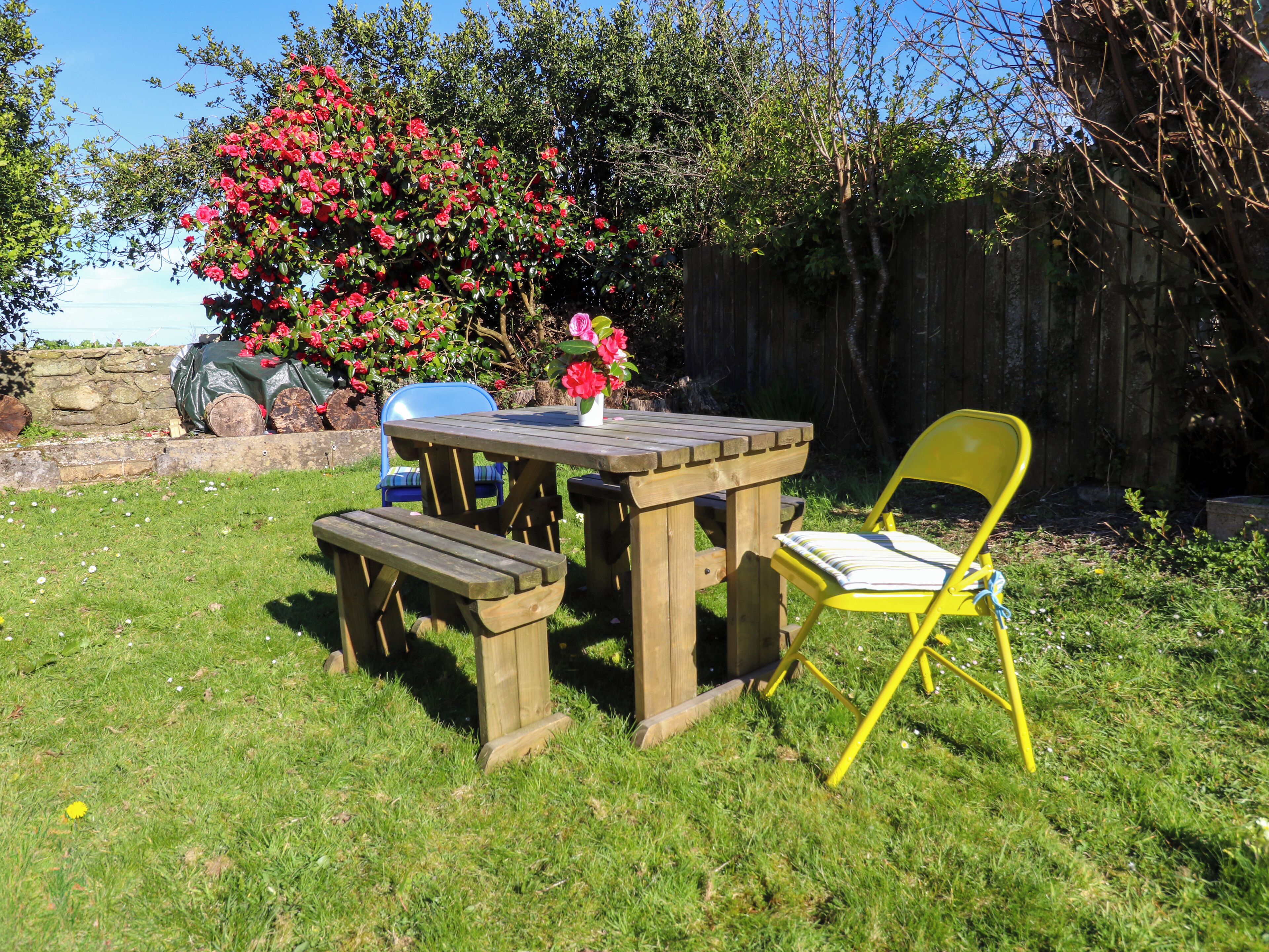 Une table de pique-nique en bois, avec des bancs et une chaise jaune, se trouve dans une cour arrière verdoyante, bordée d’un mur de pierres et agrémentée d’un arbuste en fleurs.