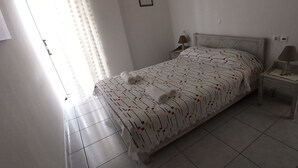 Ein ordentlich bezogenes Bett mit einem gemusterten Überwurf und zwei zusammengerollten Handtüchern, ein Nachttisch mit Lampe sowie ein Fenster mit Vorhängen.