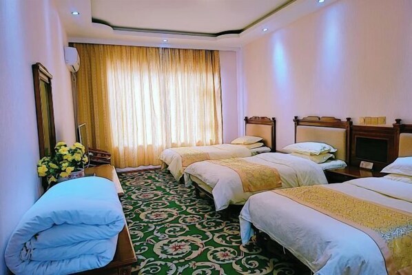 Una habitación de hotel con tres camas, alfombra verde estampada, un ventanal con cortinas y una mesita de noche con un florero.