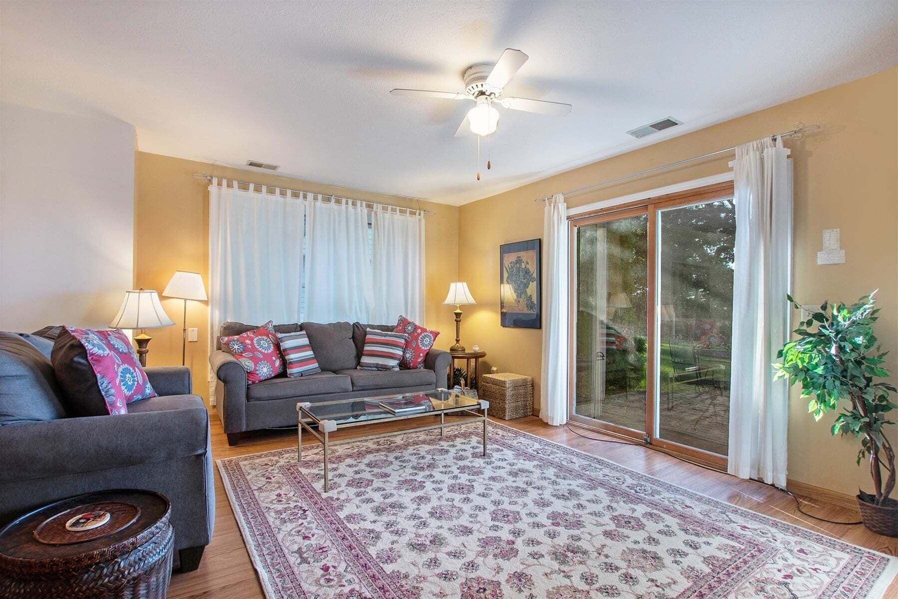 A living room with a sofa, two lamps, a coffee table, a rug, a ceiling fan, and a sliding glass door leading to a garden.