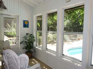 A sunlit room with large windows, wicker furniture, a potted plant, and a view of a backyard with a pool.