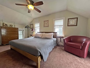 A bedroom with a bed, a red chair, a wooden dresser, a ceiling fan, and framed pictures on the walls.