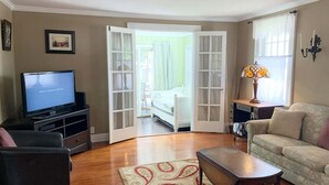 A living room with a television, a sofa, a coffee table, a patterned rug, and a bed in the adjacent room.