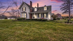 A two-story house with a front porch, a chimney, and a well-maintained lawn. - The Old Snow Farm: Lake Allegan Waterfront, Classic Farmhouse, Fire Pit, Patio (Allegan)