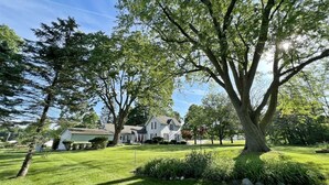A residential property with a large lawn, mature trees, and a house with a gabled roof. - The Old Snow Farm: Lake Allegan Waterfront, Classic Farmhouse, Fire Pit, Patio (Allegan)