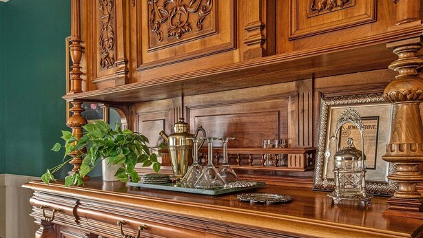 A wooden dresser with intricate carvings, a green plant, and various glassware. - The Old Snow Farm: Lake Allegan Waterfront, Classic Farmhouse, Fire Pit, Patio (Allegan)