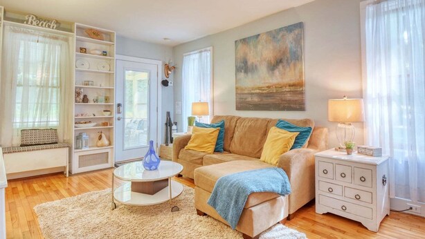 A living room with a beige sofa, a coffee table, a bookshelf, a painting, and a lamp. - Sunshine Cottage: Downtown, Screen Porch, Deck, BBQ, Walk to All Activities (Saugatuck)
