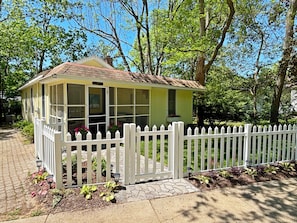 A small, single-story building with a white picket fence, a covered porch, and a brick pathway leading to the entrance.