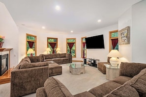 A living room with a sectional sofa, a glass coffee table, and a flat-screen TV mounted on the wall. - Great Escape: Spacious, Home Theater, Fire Pit, Walk to Downtown (Saugatuck)