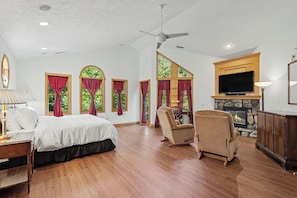 A spacious bedroom with a large bed, a sitting area with a chair, a fireplace, and multiple windows with curtains.