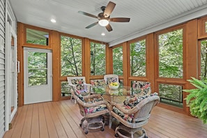A covered outdoor seating area with wicker furniture and a glass table, surrounded by large windows.