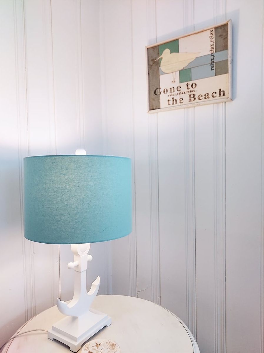 A white table with a blue lampshade and a white base, a wall with vertical paneling, and a framed sign with a bird and text.