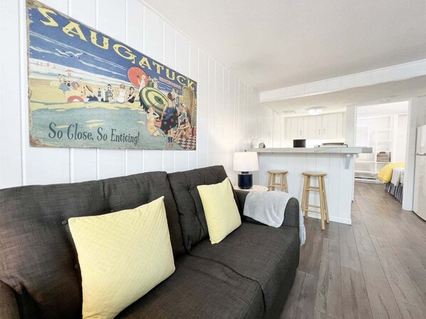 Un salon moderne avec un canapé foncé, des coussins jaunes, une cuisine en arrière-plan et une affiche vintage accrochée au mur.