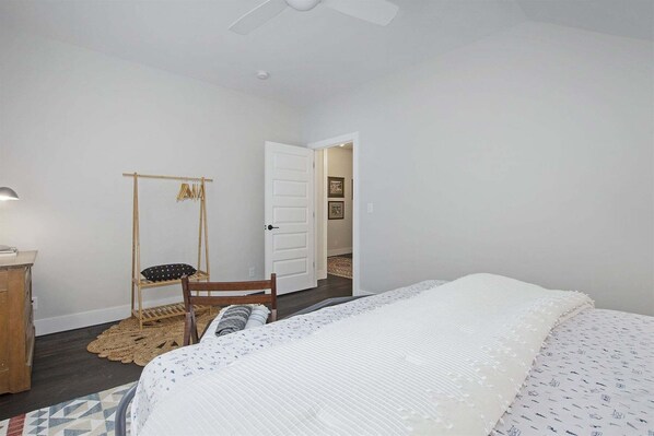 Un dormitorio con una cama, una silla de madera, una mesita, una lámpara y una puerta que da a otra habitación.
