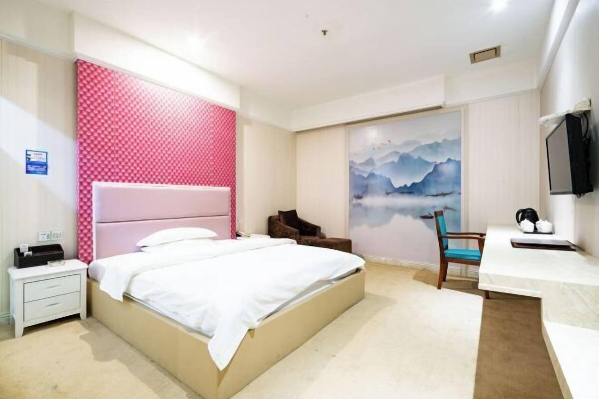A bedroom with a bed, a desk, a chair, a TV, and a wall with a mountain painting.