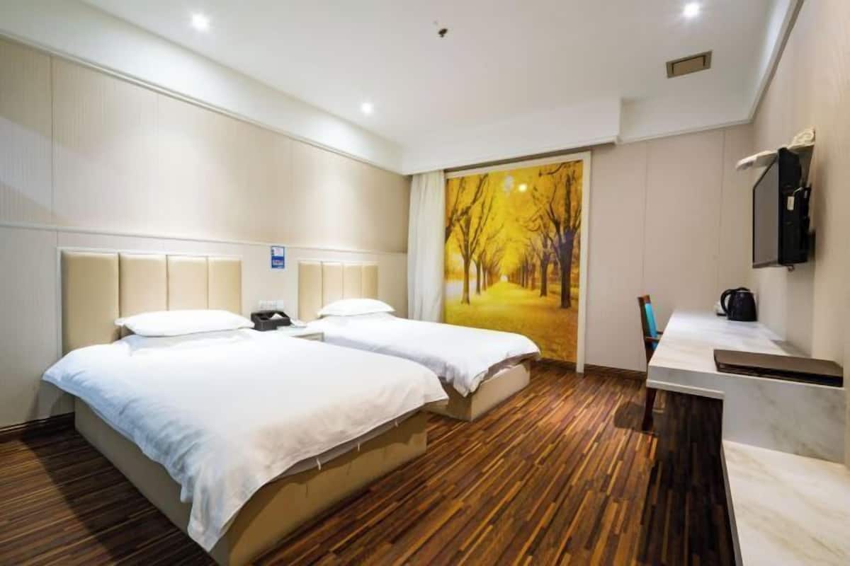 A hotel room with two beds, a wooden floor, a painting of a tree-lined path, a desk with a chair, and a mirror.
