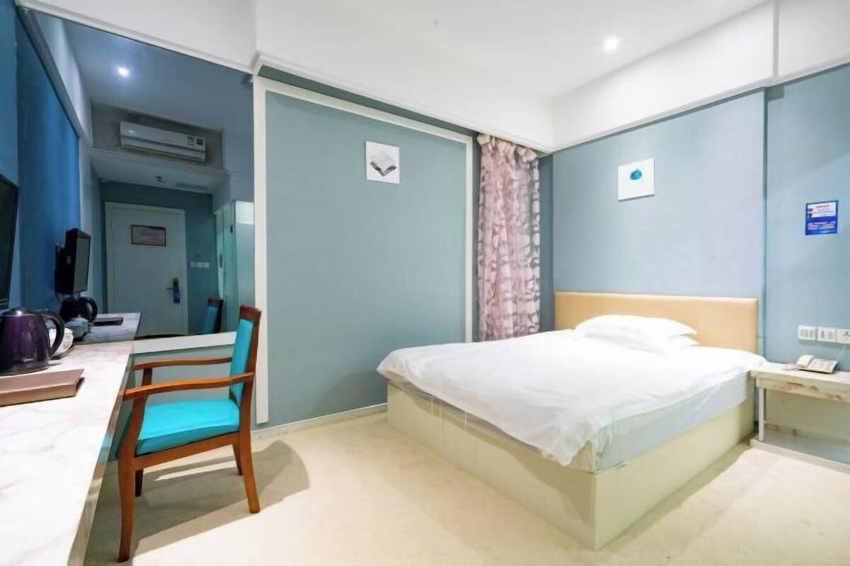 A hotel room with a bed, a desk with a chair, a nightstand, and an air conditioning unit.
