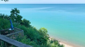 A wooden deck overlooking a beach with clear water and dense foliage. - Lagom Sunset Retreat: Hot Tub, Lake Michigan Beach, Home Theater, Sunset Views (Fennville)