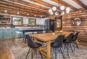 A rustic log cabin interior with a wooden dining table, black chairs, a kitchen with a refrigerator, and a large mirror on the wall.