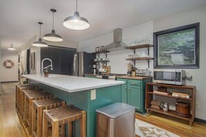 Une cuisine moderne dotée d’un îlot central, de tabourets de bar, d’une poubelle en acier inoxydable et d’une fenêtre donnant sur un espace verdoyant.