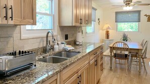 A kitchen with wooden cabinets, granite countertops, a built-in oven, and a dining area with a table and chairs. - Bevlyn Cottage: Walk to Lake Michigan, Charming Cottage w/ Deck & Fire Pit (Douglas)