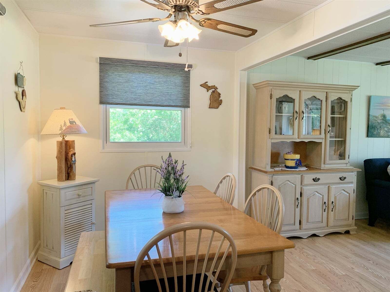 A dining area with a wooden table, chairs, a ceiling fan, a window with a view, and a cabinet with glass doors.