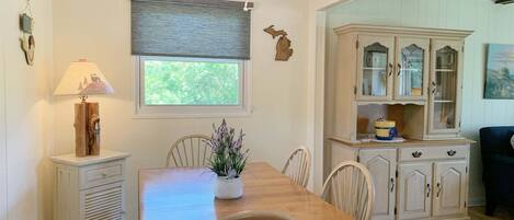 A dining area with a wooden table, chairs, a ceiling fan, a window with a view, and a cabinet with glass doors.