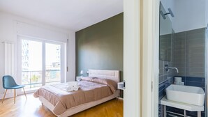 A modern bedroom with a bed, a chair, a bathroom with a sink, and a view through a glass door. - Chiaia Mon Amour Elegant Flat by Napoliapartments (Naples)