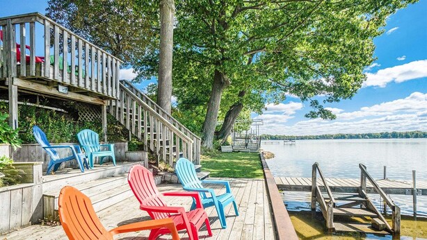 Una terraza de madera con sillas Adirondack, una escalera y un muelle que se adentra en un lago.