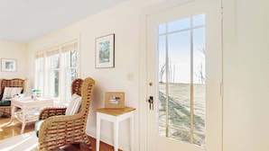 A room with a large window, a wicker chair, a small table, and a door leading outside. - Summer Haven: Lake Michigan Views, Fire Pit, BBQ, Classic Cottage (Fennville)