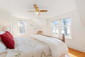 Une chambre à coucher avec un lit, un ventilateur de plafond, deux fenêtres, une chaise et une lampe.