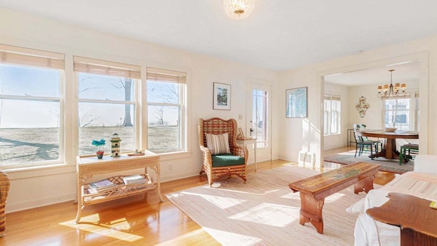 A spacious living room with large windows, a wooden bench, a wicker chair, and a dining area with a table and chairs. - Summer Haven: Lake Michigan Views, Fire Pit, BBQ, Classic Cottage (Fennville)