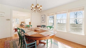 A dining room with a wooden table, green chairs, a chandelier, and large windows with a view of trees. - Summer Haven: Lake Michigan Views, Fire Pit, BBQ, Classic Cottage (Fennville)