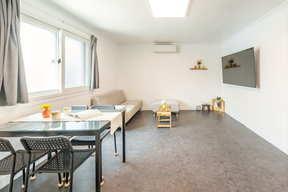 A modern, minimalist living room with a dining area, a sofa, a small table with a vase, and a bed in the adjacent room.
