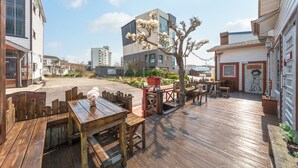 A wooden deck with tables and chairs, surrounded by buildings and a tree with white flowers. - Sokcho Seohyeon Ine Pension (Sokcho)